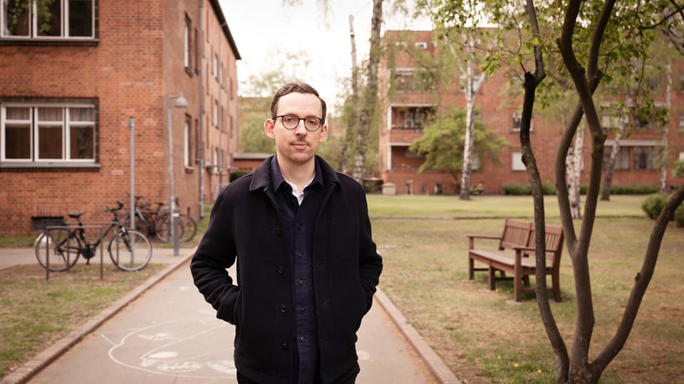 Eine Person in dunkler Jacke steht auf einem gepflasterten Weg zwischen roten Backsteingebäuden; links lehnen Fahrräder an einem Zaun, rechts stehen Bäume und eine Parkbank auf einer Grünfläche.