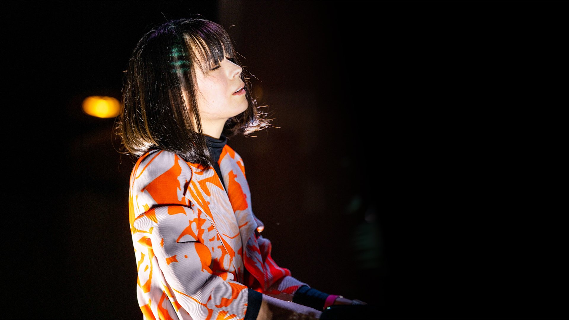 Bühnenaufnahme von Alice Sara Ott am Klavier: Seitliche Ansicht einer Person beim Pianospiel auf einer abgedunkelten Bühne. Gemusterte Jacke in Orange‑, Grau‑ und Schwarztönen, kurze dunkle Haare. Warmes Spotlicht beleuchtet Oberkörper und Instrument, der Hintergrund bleibt dunkel. Konzertmotiv zur klassischen Live‑Aufführung mit Orchester.