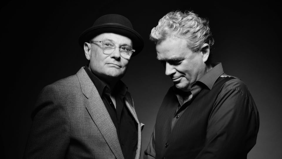 Schwarzweiß-Pressefoto zu „Pigor & Eichhorn – Grundkurs II“ mit zwei Männern vor dunklem Hintergrund, einer mit Hut und Brille, der andere mit Weste, beide nebeneinander stehend.