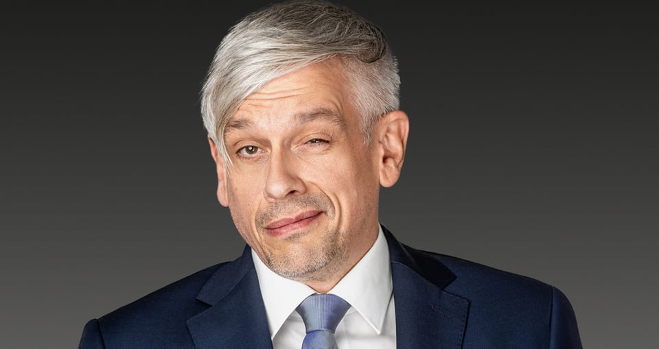 Mann zwinkernd in blauem Sakko und grauem Hintergrund