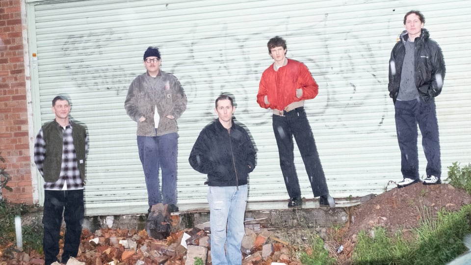 Bandfoto von Basement mit fünf Personen vor einer hellgrünen Rolltorwand auf einem Industriegelände, verteilt zwischen Bauschutt und Grasflächen, darunter eine Person in roter Jacke