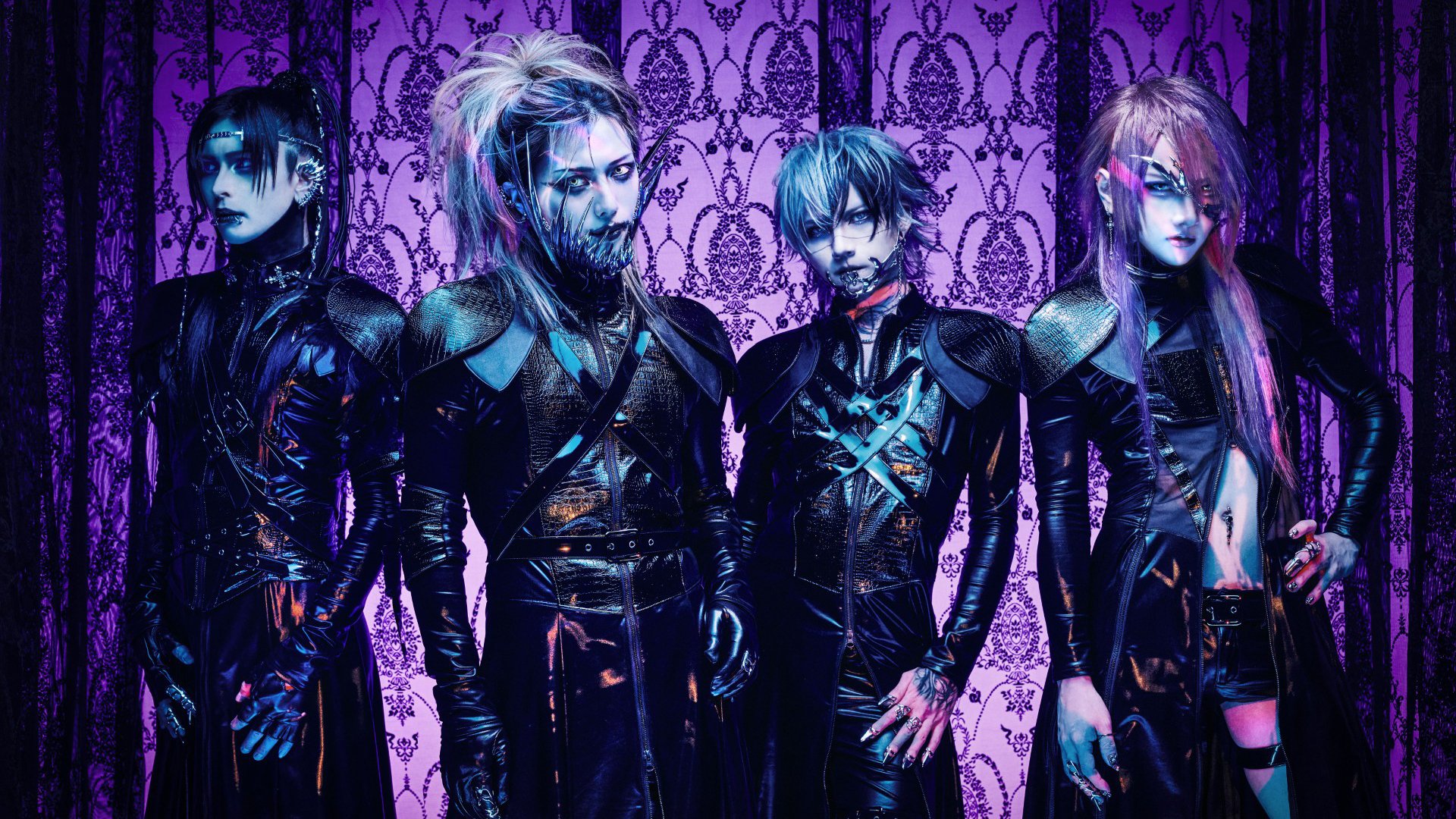 Vier Personen stark geschminkt in schwarzen, futuristischen Outfits mit auffälligen Frisuren und Metall-Accessoires vor violett gemusterter Wand mit Ornamenten in blau‑lila Licht