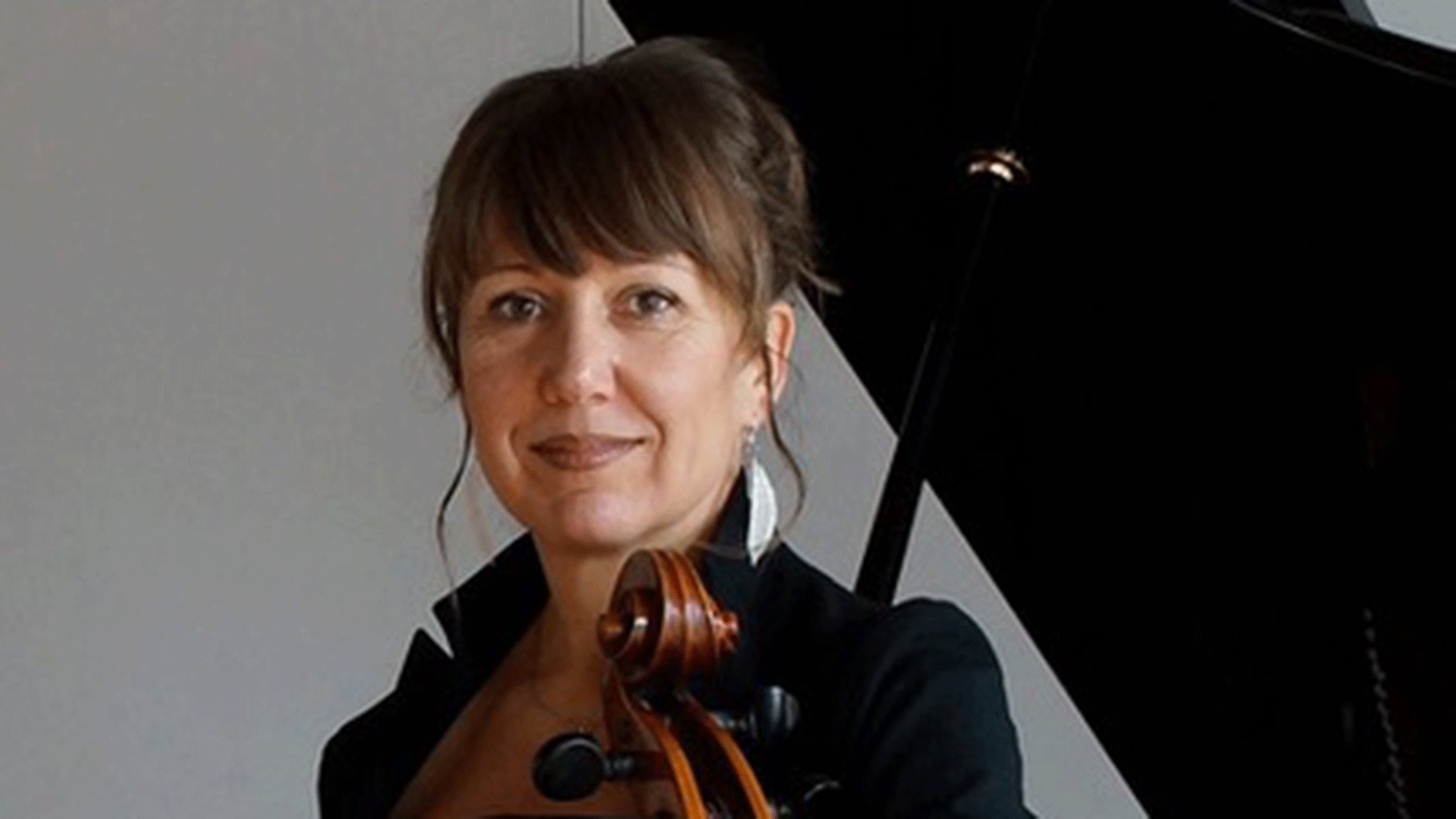 Porträt einer Cellistin vor einem Flügel. Die Person mit hochgestecktem Haar und schwarzer Konzertkleidung hält ein Violoncello vor sich und blickt in die Kamera.