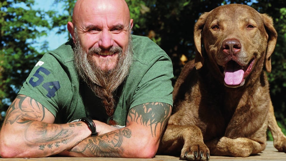 Holger Schüler in grünem T-Shirt auf die Ellbogen gestützt blickt lachend in die Kamera, neben ihm ein Hund