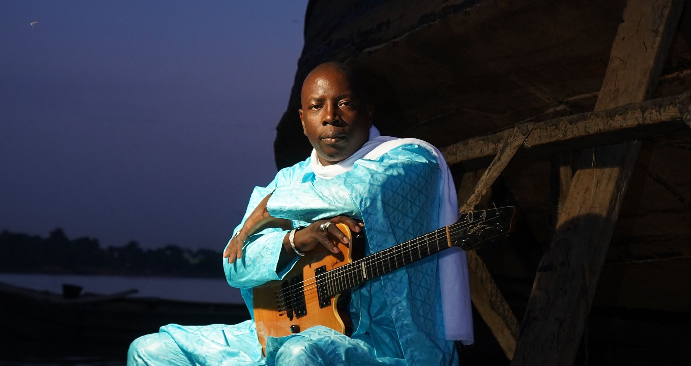 Foto des Sängers „Vieux Farka Toure“, sitzend mit einer Gitarre vor einem abendlichen Hintergrund im Freien