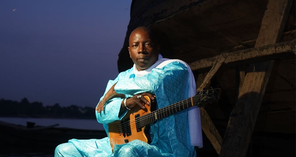 Foto des Sängers „Vieux Farka Toure“, sitzend mit einer Gitarre vor einem abendlichen Hintergrund im Freien