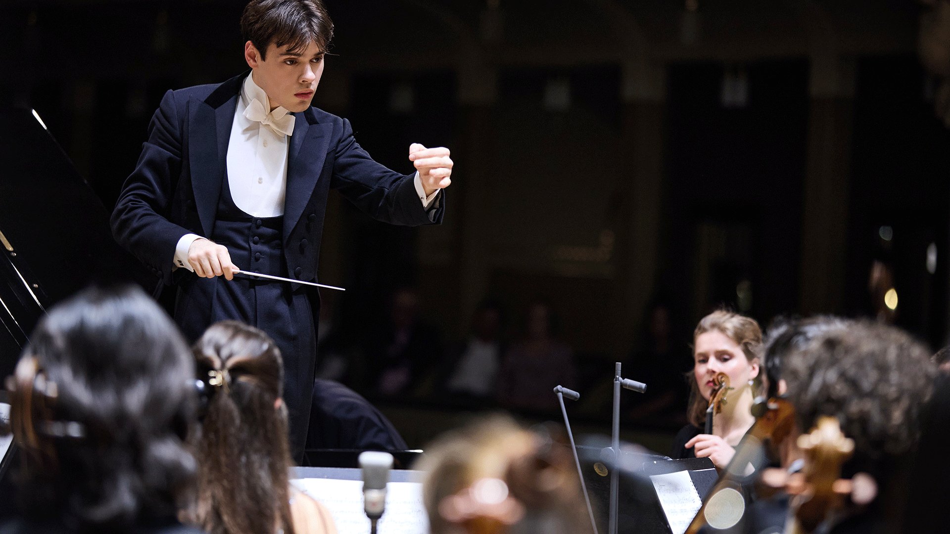 Person auf dem Dirigentenpodest während eines Konzerts des Münchner Festspielorchesters mit erhobenem Taktstock vor den Musikerinnen und Musikern des Orchesters