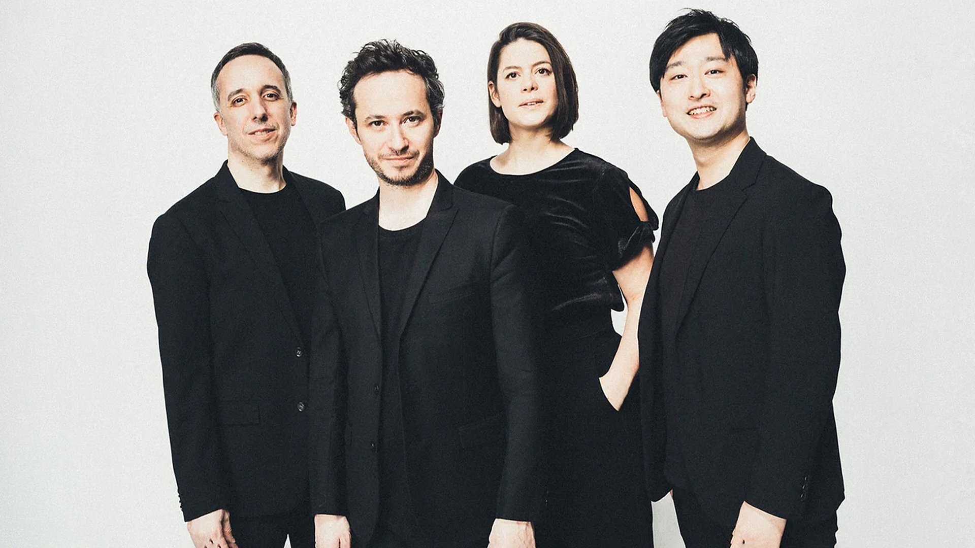 Quatuor Ébène mit vier Musiker:innen in schwarzer Kleidung vor hellem Hintergrund, nebeneinander stehend in Studioaufnahme ohne sichtbare Instrumente.