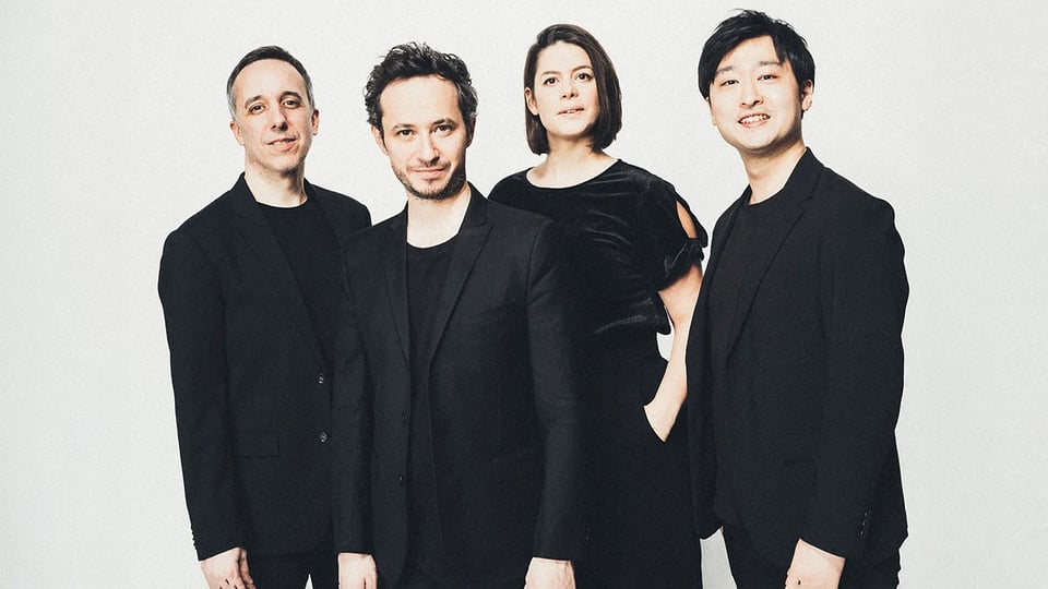 Quatuor Ébène mit vier Musiker:innen in schwarzer Kleidung vor hellem Hintergrund, nebeneinander stehend in Studioaufnahme ohne sichtbare Instrumente.