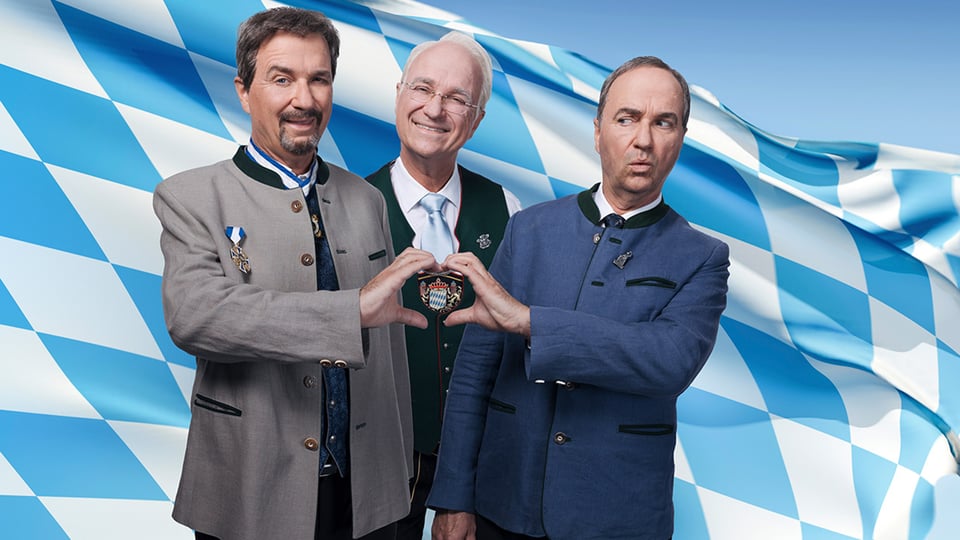 Wolfgang Krebs und zwei weitere Personen stehen vor einer Bayern-Flagge und halten die Hände zu einer Herzform zusammen