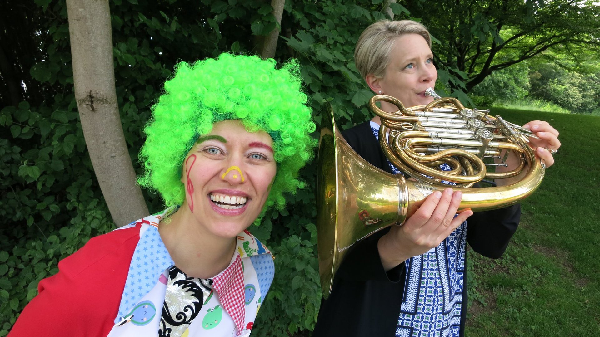 Zwei Frauen im Grünen: Eine lächelt mit grüner Clown-Perücke und bunter Gesichtsbemalung in die Kamera, die andere spielt ein Horninstrument.
