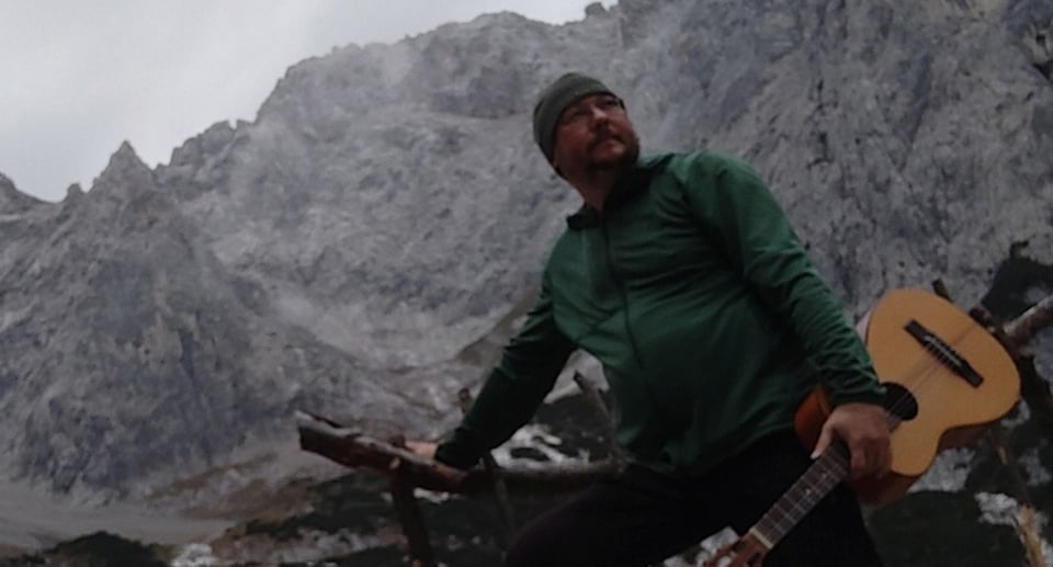 Mann mit Mütze und Gitarre vor gebirgigem Hintergrund