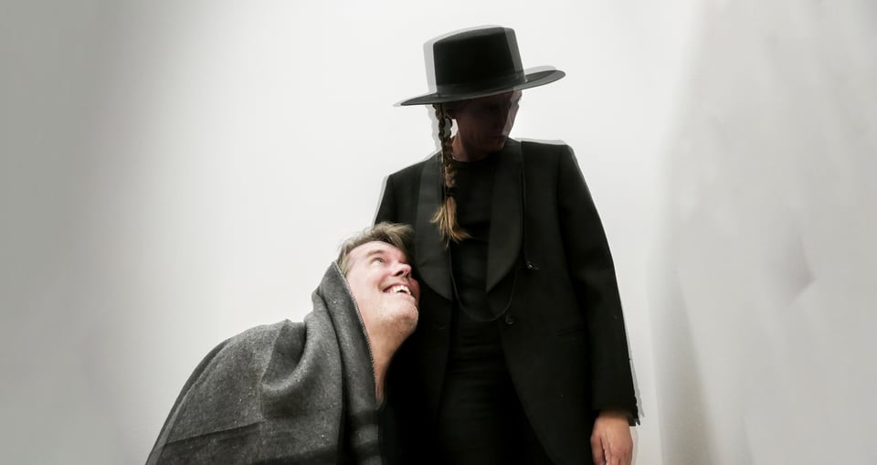 Eine schwarz gekleidete Person mit Zopf und schwarzem Hut und eine Person in eine Decke gehüllt 