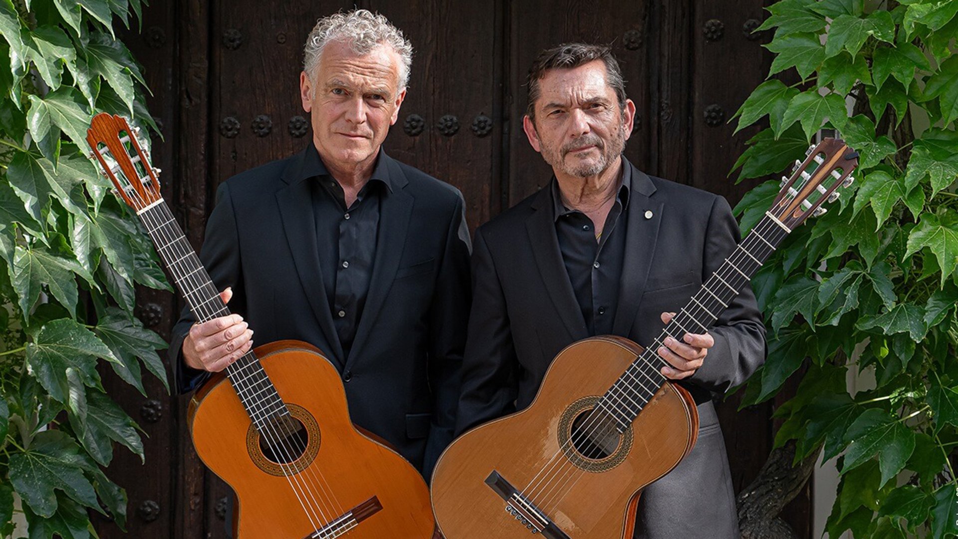 Das Gitarrenduo Wiedermann und GAllardo del Rey mit Gitarren vor einer Holztür, links und rechts im Bild Kletterpflanzen