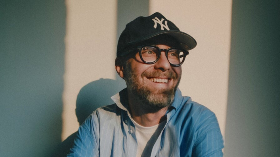 Mark Forster in einem blauen Hemd mit Brille und schwarzer Kappe mit weißem Logo blick zur Seite, im Hintergrund Lichtstrahlen und Schatten