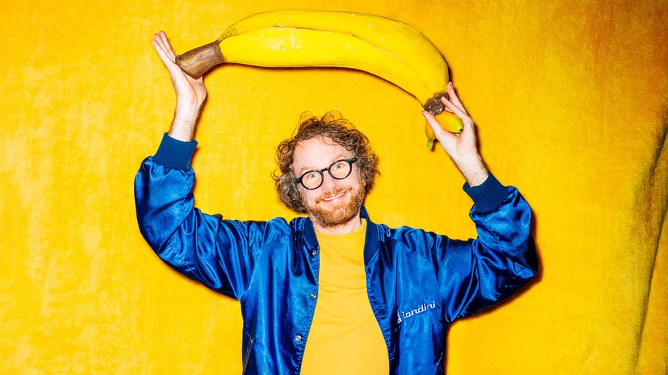 Friedemann Weise lacht vor einem gelben Hintergrund. Er hält eine große XXL Banane über dem Kopf.