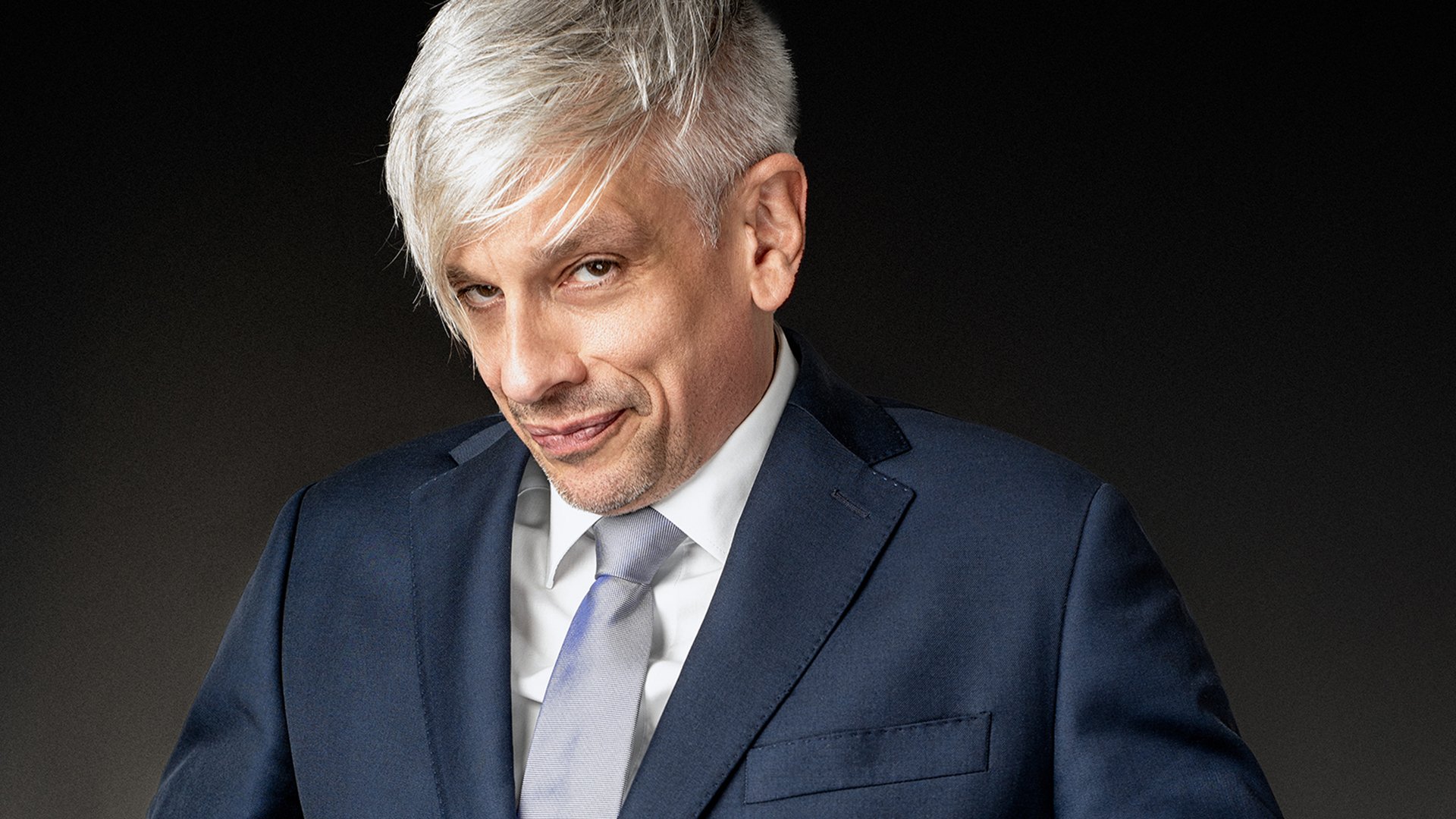 Porträt einer Person mit kurzem, hellem Haar im Anzug und Krawatte vor dunklem Hintergrund, Nahaufnahme, Pressebild für ein Kabarett- oder Comedyprogramm.