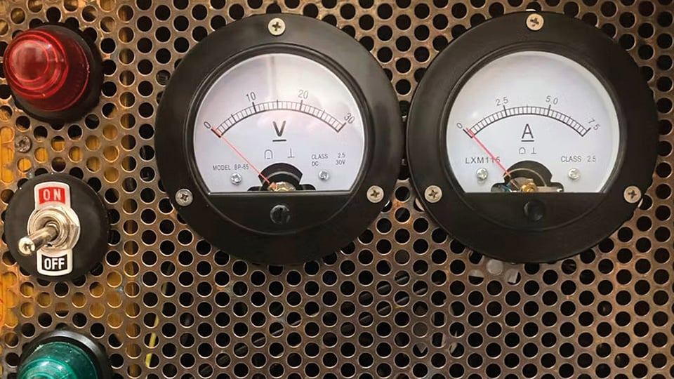 Nahaufnahme eines technischen Bedienfelds mit zwei analogen Messinstrumenten für Spannung und Stromstärke, einem roten und grünen Kontrolllicht sowie einem Kippschalter mit den Beschriftungen ON und OFF vor perforierter Metallfläche.