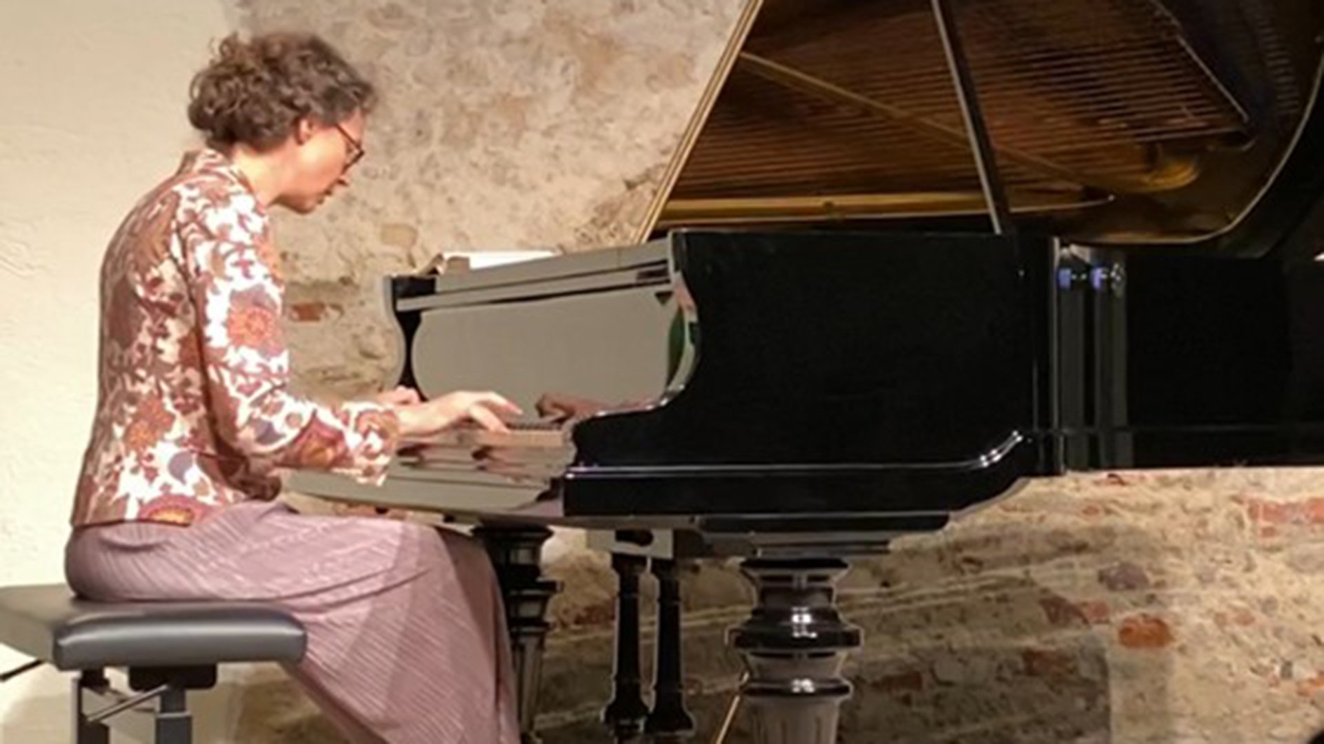 Person spielt an einem geöffneten Konzertflügel Klavier vor sitzendem Publikum in einem Saal mit Steinwand.