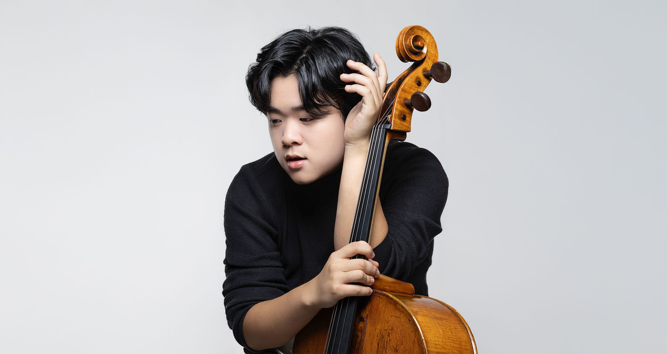 Mann in schwarzer Kledung mit Violoncello, sitzend vor hellem Hintergrund