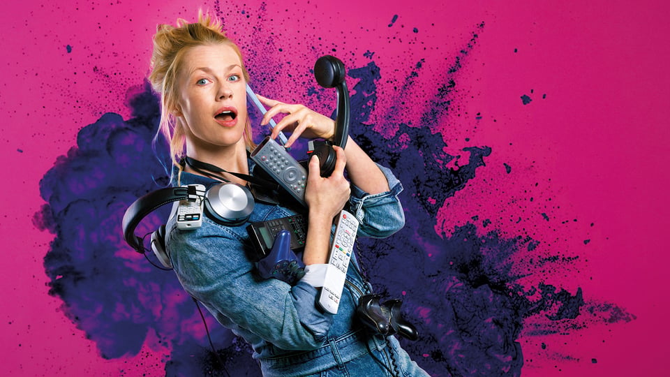 Christine Eixenberger im Jeansoutfit, die zahlreiche Alltagsgeräte wie Telefon Kopfhörer und Fernbedienungen hält, vor pink violettem Hintergrund mit Farbspritzern