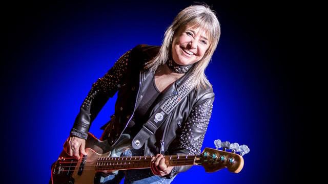 Suzi Quatro mit E‑Gitarre steht auf einer Bühne vor blauem Hintergrund.