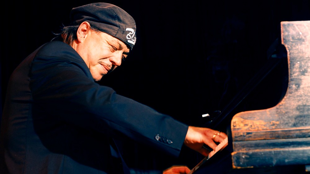 Pianist mit dunkler Kleidung und Kopftuch spielt konzentriert auf einem Flügel auf einer Bühne, Nahaufnahme von der Seite mit sichtbaren Händen auf den Tasten und dunklem Hintergrund.