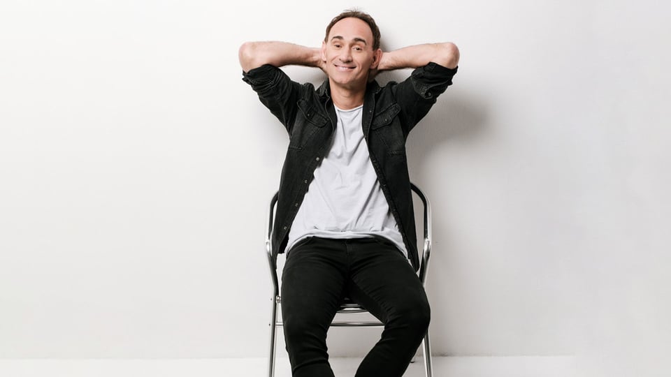 Porträt des Kabarettisten Gernot Kulis auf einem Stuhl sitzend vor hellem Hintergrund, Werbefoto zum Comedy-Programm von Gernot Kulis.