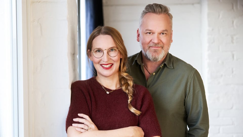 Franca Cerutti in dunkelrotem Pulli und Christian Weiss in grünem Hemd posieren vor einer weißen Mauer