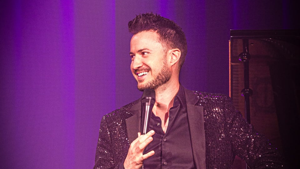 Foto für das Event Florian Wagner – The Flo Must Go On mit dem Künstler, der ein Mikrofon in der Hand hält, vor einem lilafarbenen Hintergrund