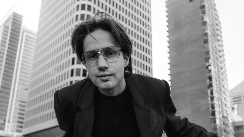 Schwarz-weißes Porträt von Jude York mit Brille und mittellangen Haaren, die sich im Vordergrund leicht nach vorne lehnt; im Hintergrund sind hohe Gebäude einer Stadt zu sehen.