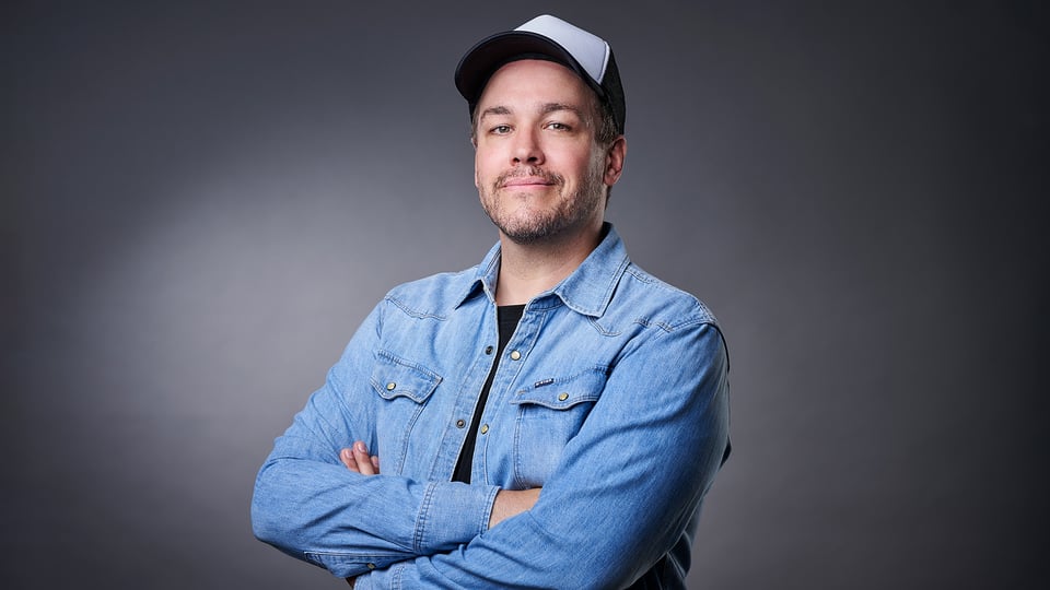 Jan von Weyde steht mit Jeanshemd und weißer Cap vor einem grauen Hintergrund und schaut in die Kamera, während er seine Arme verschränkt.