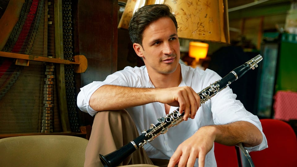 Promobild für das Event ‚Concertgebouw Chamber Orchestra / Ottensamer‘: Eine Person sitzt in informeller Haltung auf einem Stuhl und hält eine Klarinette in den Händen. Im Hintergrund sind verschiedene Möbelstücke, eine Lampe und Teile eines geöffneten Klaviers sichtbar