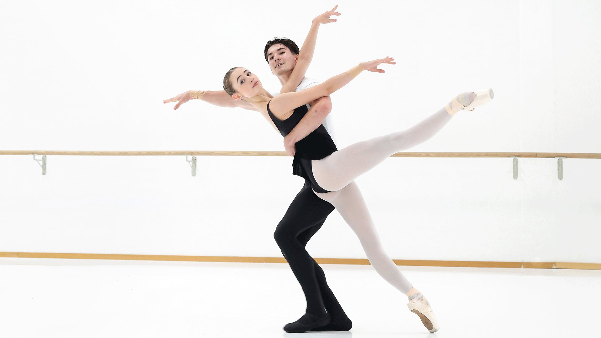 Ballettpaar in einem hellen Tanzsaal vor einer Ballettstange. Eine Person in Spitzenschuhen streckt ein Bein waagerecht aus, während die andere Person die Figur hebt und beide die Arme elegant ausbreiten.