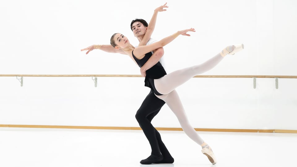 Ballettpaar in einem hellen Tanzsaal vor einer Ballettstange. Eine Person in Spitzenschuhen streckt ein Bein waagerecht aus, während die andere Person die Figur hebt und beide die Arme elegant ausbreiten.