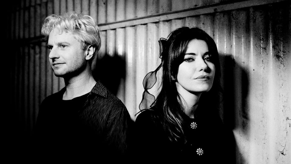 Schwarz‑weiß‑Foto des Duos Blood Red Shoes mit zwei Personen, die nebeneinander vor einer geriffelten Metallwand stehen; linke Person mit hellem Haar und dunkler Kleidung, rechte Person mit längerem Haar und dunkler Kleidung mit zwei dekorativen Ansteckern