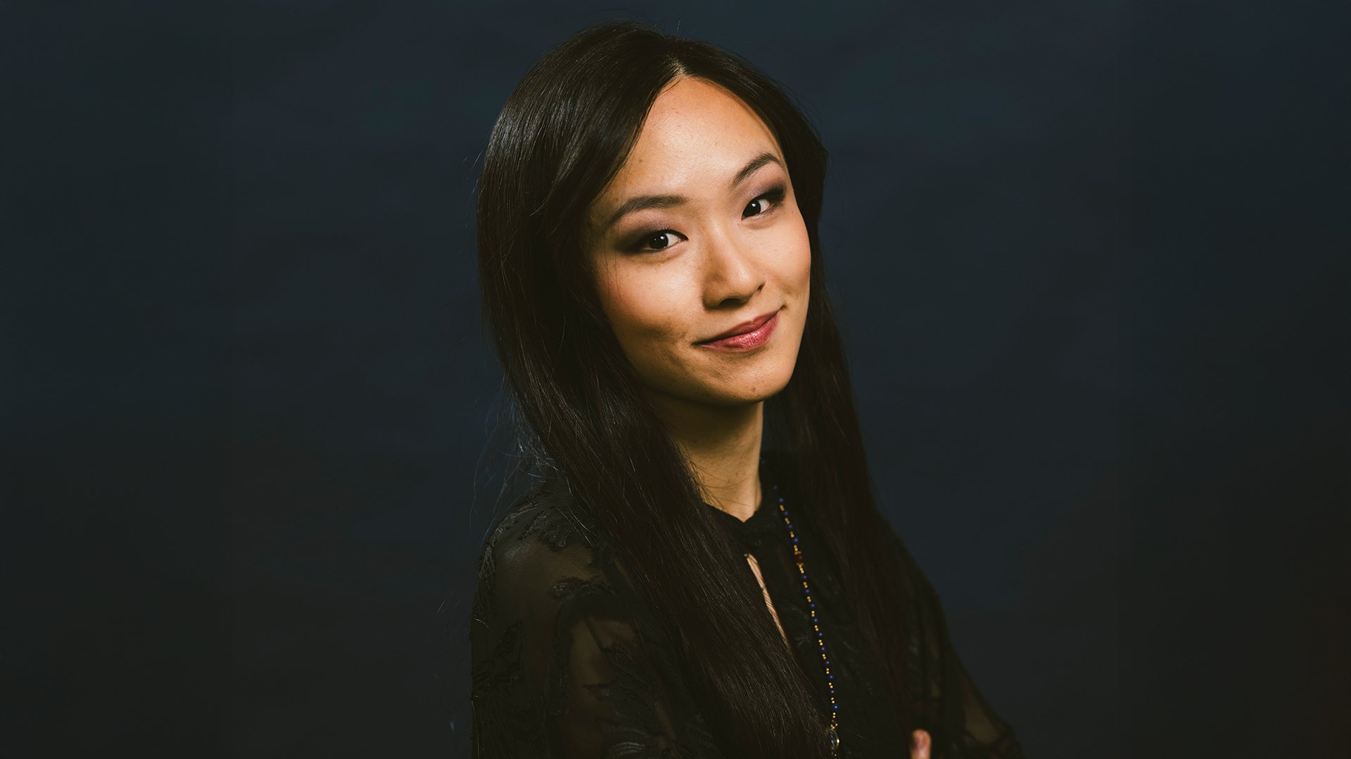 Ein Portrait von Liya Wang mit verschränkten Armen in schwarzer Kleidung vor dunklem Hintergrund