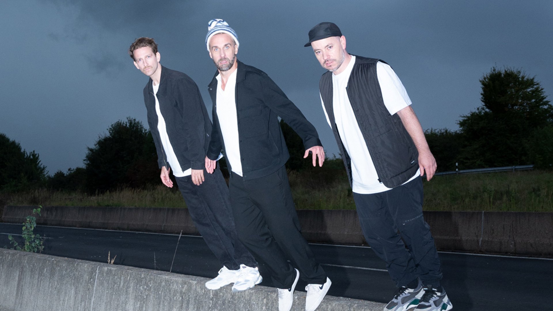 OK KID Bandfoto: Drei Männer stehen leicht nach vorne geneigt auf einer Betonmauer im Freien bei Dämmerung, tragen dunkle Kleidung und schauen in die Kamera, Straße und Bäume im Hintergrund.