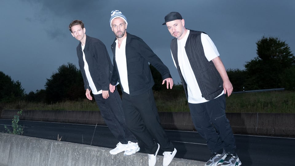 OK KID Bandfoto: Drei Männer stehen leicht nach vorne geneigt auf einer Betonmauer im Freien bei Dämmerung, tragen dunkle Kleidung und schauen in die Kamera, Straße und Bäume im Hintergrund.