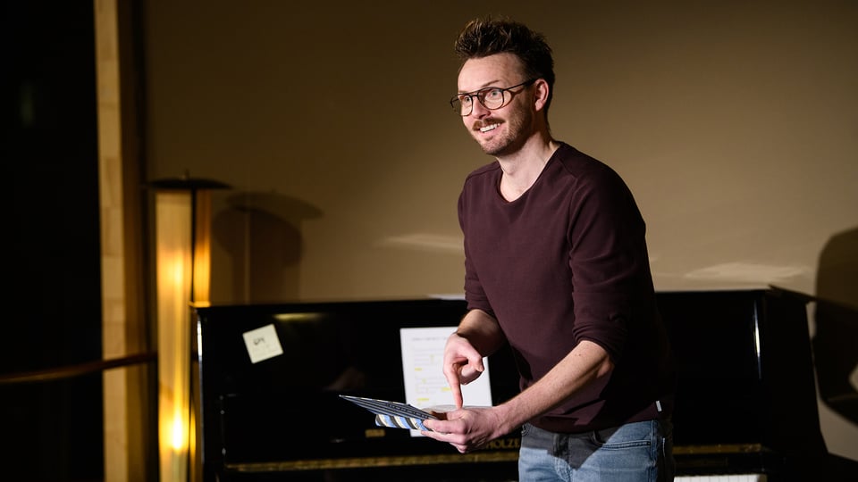 Ein Foto für die Veranstaltung ‚All das Schöne‘ mit dem Künstler, der etwas in der Hand hält und darauf zeigt, im Hintergrund ein Klavier