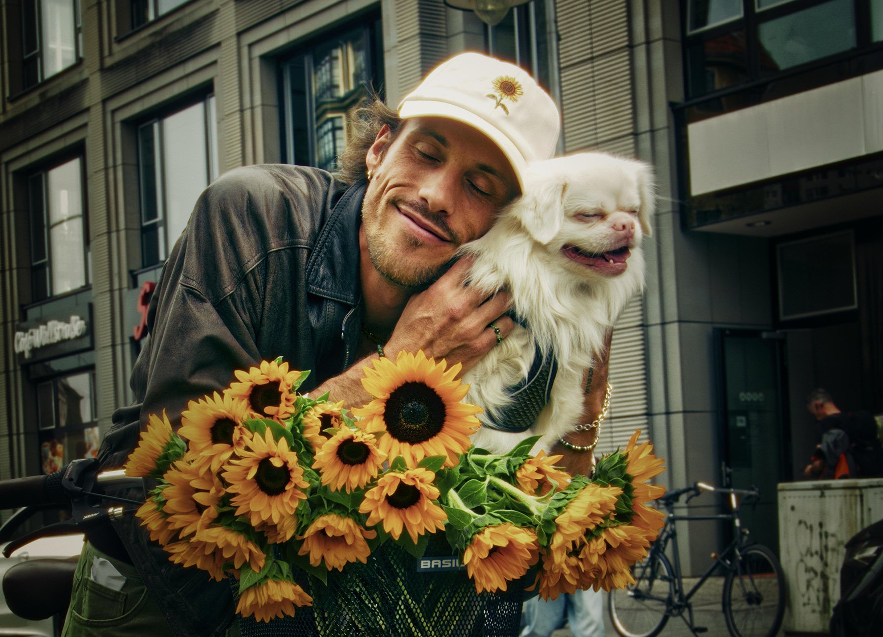 Mann mit weisser Kappe vor Häuserwand mit weissem Hund und einem Strauß Sonnenblumen