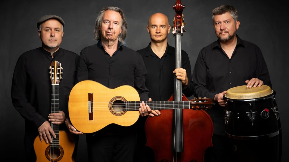 Vier Musiker stehen nebeneinander vor dunklem Hintergrund, tragen schwarze Kleidung und halten Instrumente wie zwei Gitarren, einen Kontrabass und eine Handtrommel in einer Studioaufnahme.