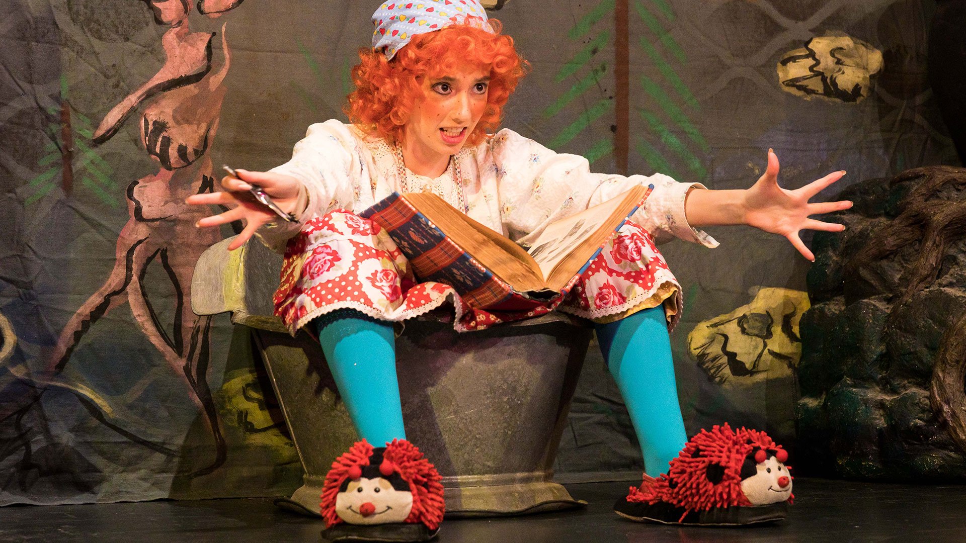 Foto für das Event Die kleine Hexe, mit einer Frau, die spielt und verkleidet ist, in einem Würfel sitzt und ein Buch sowie Marienkäfer‑Hausschuhe hat.