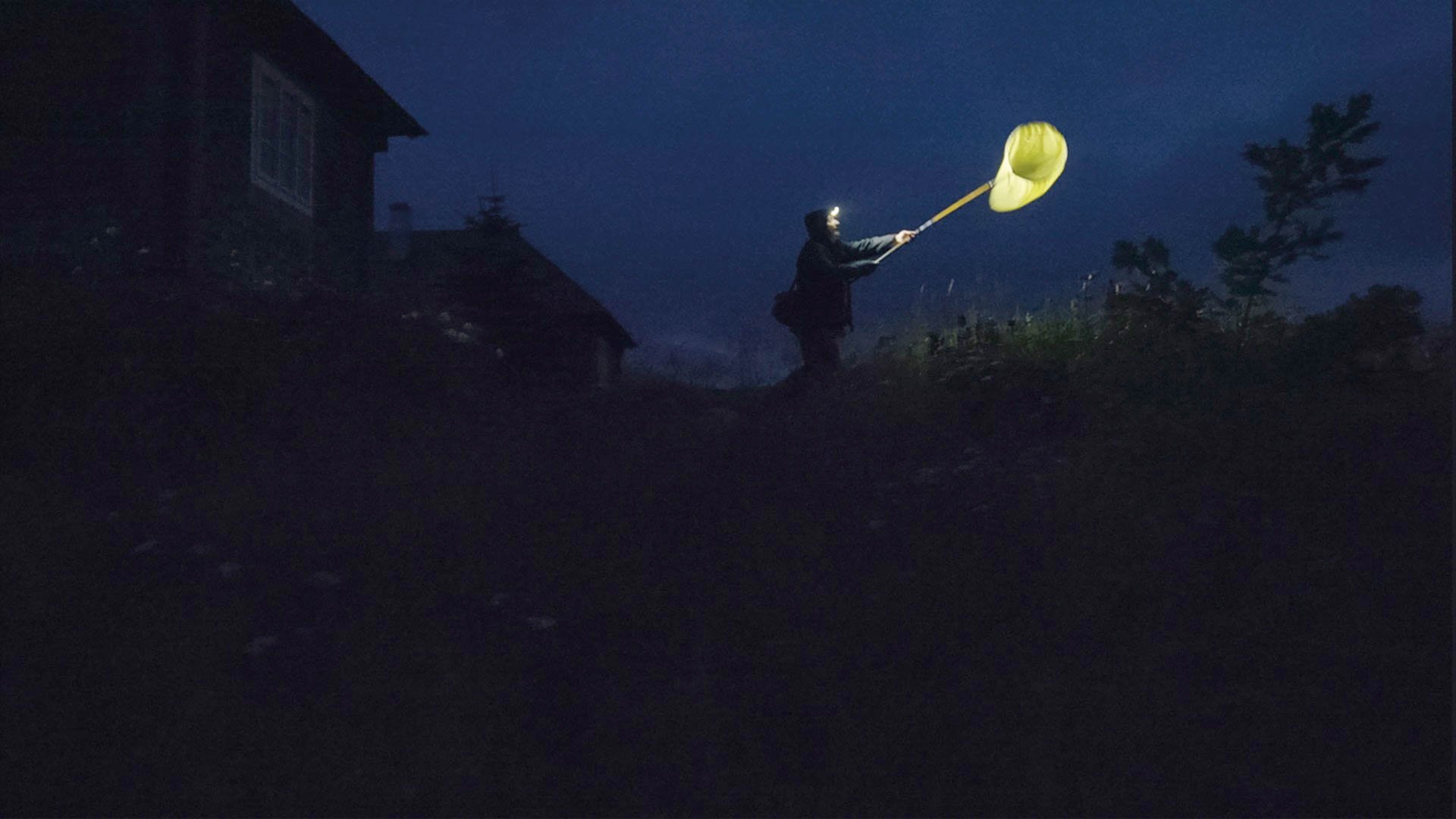 In einer nächtlichen Landschaft steht eine Person auf einer Wiese neben einem Haus und hält mit ausgestrecktem Arm ein leuchtend gelbes, mondähnliches Objekt; der Himmel ist dunkelblau, und die Szene wird vom starken Kontrast zwischen Dunkelheit und dem hellen Licht des Objekts bestimmt.