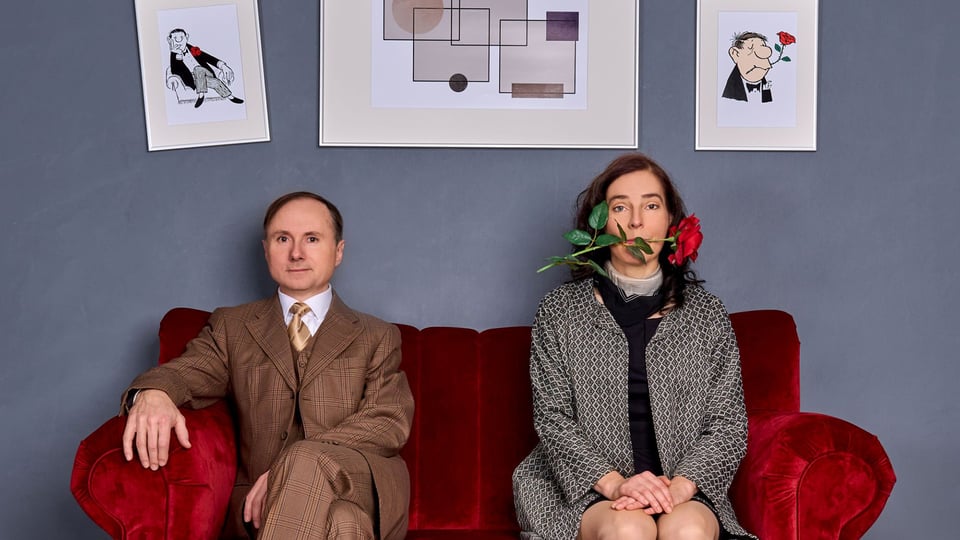 Zwei Personen sitzen auf einem roten Sofa vor grauer Wand mit gerahmten Illustrationen, eine Person im Anzug links, eine Person rechts mit roter Rose im Mund, beide frontal zur Kamera.