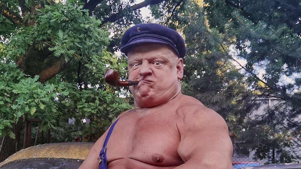 Muskulöser Mann mit Mütze und Pfeife posiert oberkörperfrei im Freien, blickt mit grimmigem Gesichtsausdruck in die Kamera, Bäume im Hintergrund