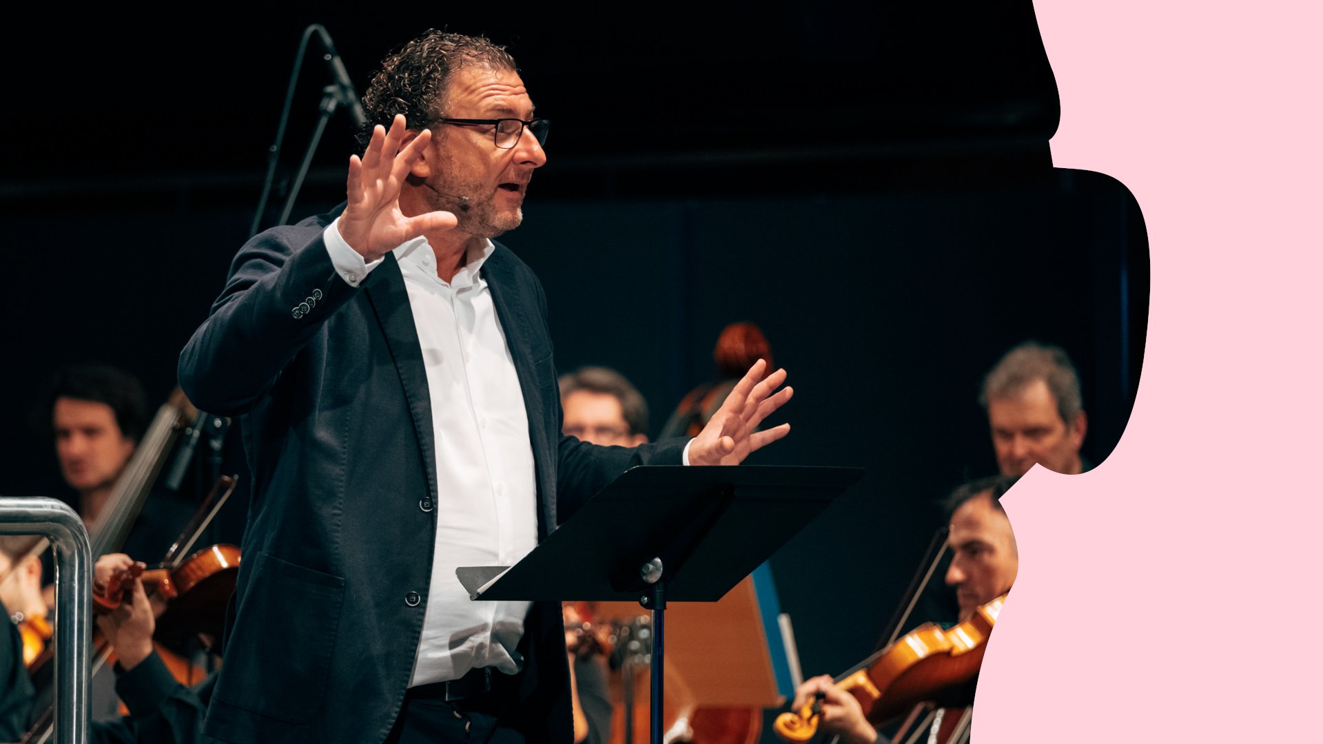 Person am Rednerpult vor einem Orchester auf einer Konzertbühne, umgeben von rosafarbenen grafischen Elementen am Bildrand.
