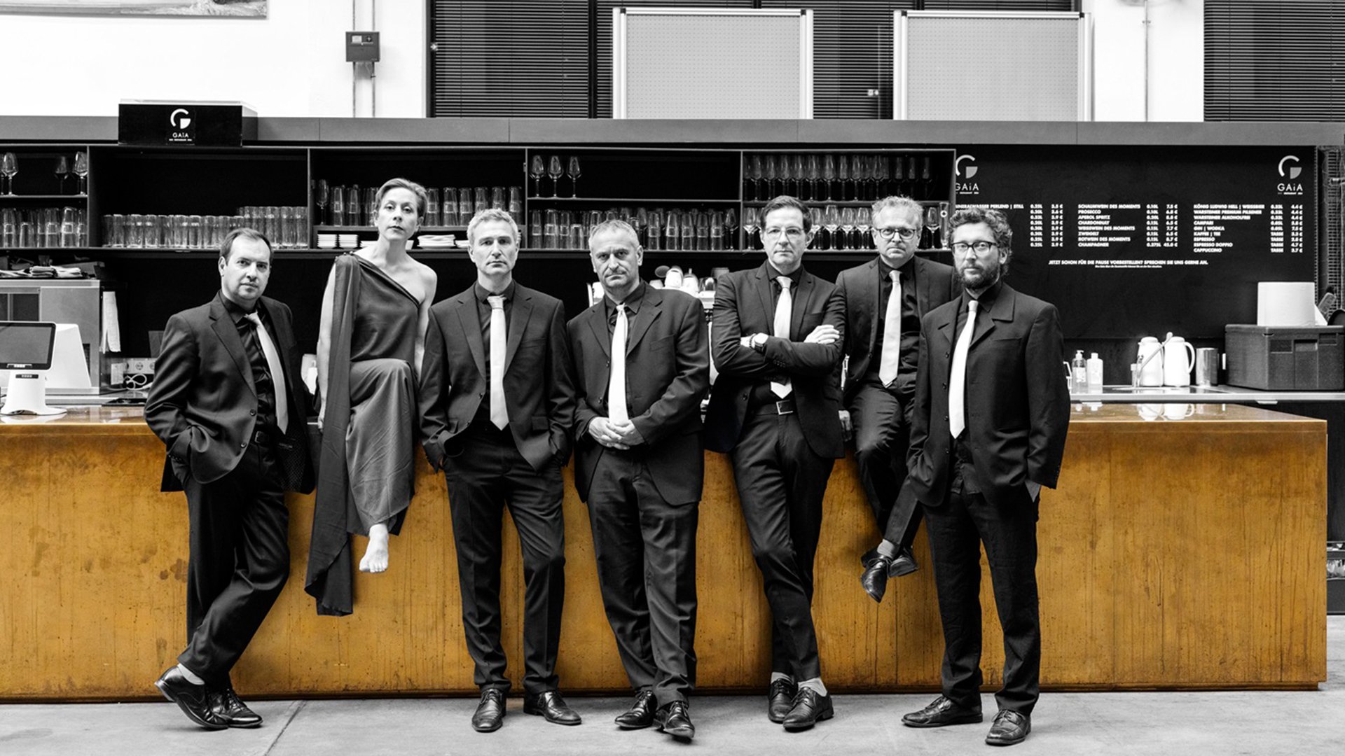 Bandfoto von Goldmund mit sieben Personen in dunkler Konzertkleidung vor einer Bar, sechs in Anzügen mit hellen Krawatten und eine Person in langem asymmetrischem Kleid, vor Regalen mit Gläsern und Getränkekarte im Hintergrund
