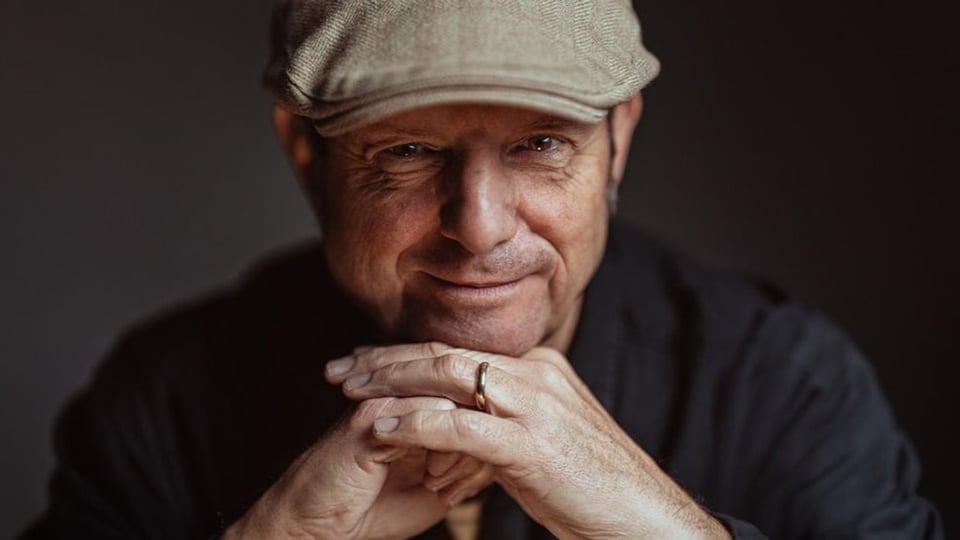 Porträt von Ludwig Seuss mit Mütze, der die Hände gefaltet vor sich hält, vor dunklem Hintergrund.