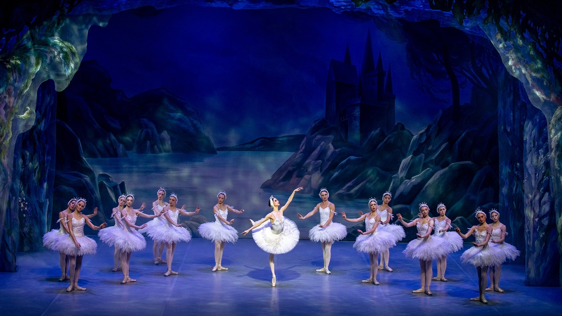 Bühnenfoto aus Schwanensee mit einer Solotänzerin in weißem Tutu im Zentrum, umgeben von weiteren Balletttänzerinnen in weißen Kostümen auf einer blau beleuchteten Bühne vor einer nächtlichen Seen- und Schlosskulisse